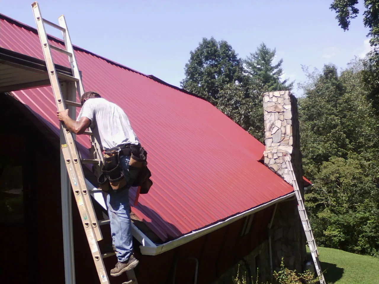 Roofer on a ladder beside a red metal roof and stone chimney — Soffit & Fascia Repair by Roofing Brothers in Sanford